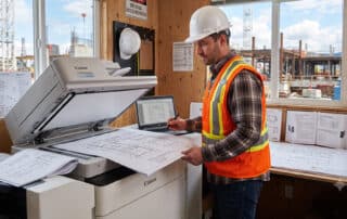 Construction project manager reviewing printed wide-format blueprints next to a high-speed office multifunction copier